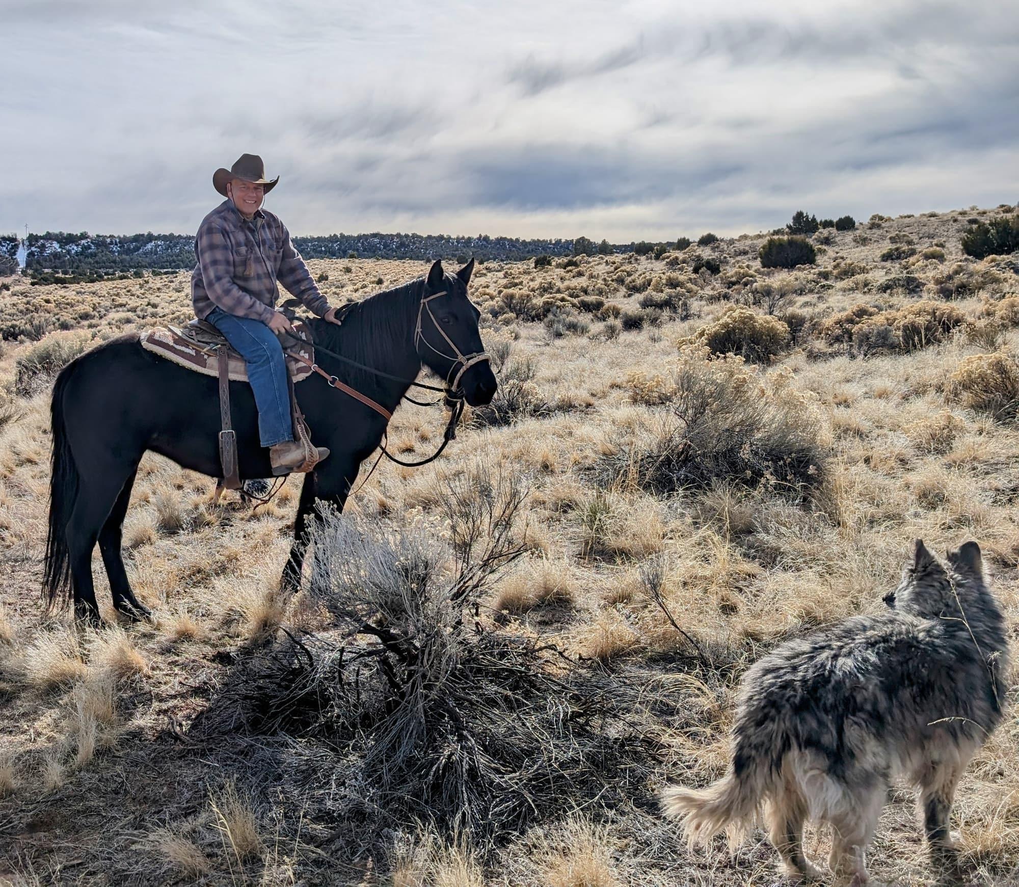 Paul D'Souza riding a black quarter horse across winter sagebrush, a shepherd dog walking alongside under a heavy grey sky
